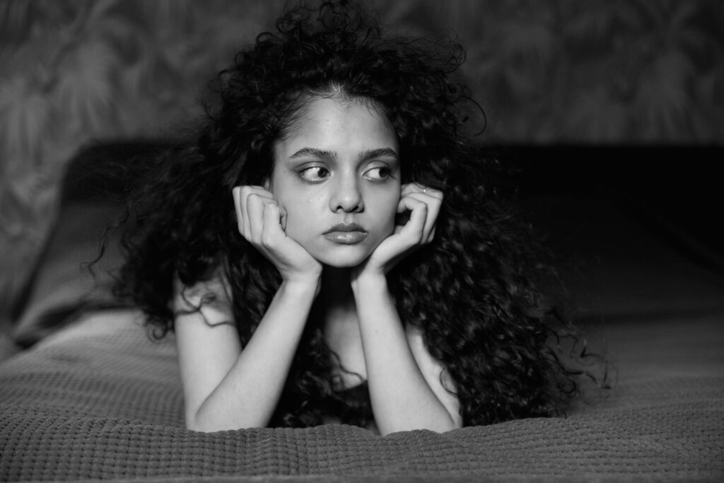 A thoughtful young woman with curly hair lies on a bed in a black and white portrait. Moody and introspective.